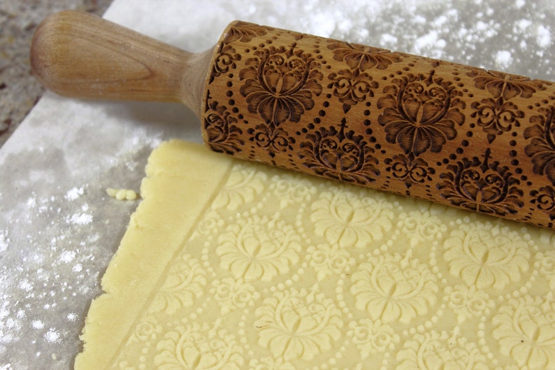 May include: A wooden rolling pin with an embossed floral pattern rests on a sheet of dough, creating a matching design. The rolling pin is light brown, and the dough is a pale yellow. Flour dusts the surface.