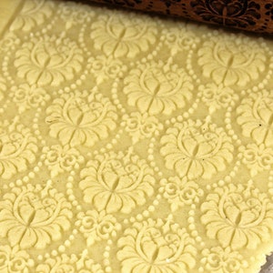 May include: Close-up of a light yellow dough with an embossed floral pattern. A wooden rolling pin with a matching floral design is partially visible at the top. The pattern includes large flowers and dotted lines.