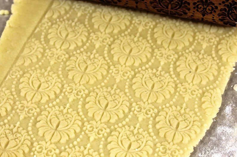 May include: Close-up of a light yellow dough with an embossed floral pattern. A wooden rolling pin with a matching floral design is partially visible at the top. The pattern includes large flowers and dotted lines.