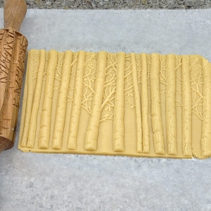 May include: A wooden rolling pin with a tree bark design is used to roll out a sheet of dough with a tree bark pattern.