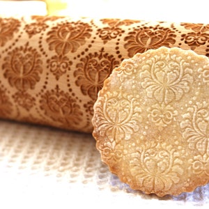 May include: A close-up of a wooden rolling pin with an intricate floral pattern. The rolling pin is used to create decorative patterns on dough. A cookie with the same pattern is shown next to the rolling pin.