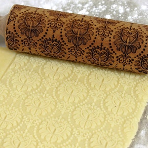 May include: A wooden rolling pin with an ornate floral pattern is pressed into a sheet of dough, creating an embossed design. The rolling pin is light brown, and the dough is a pale yellow color. Flour is sprinkled around the dough.
