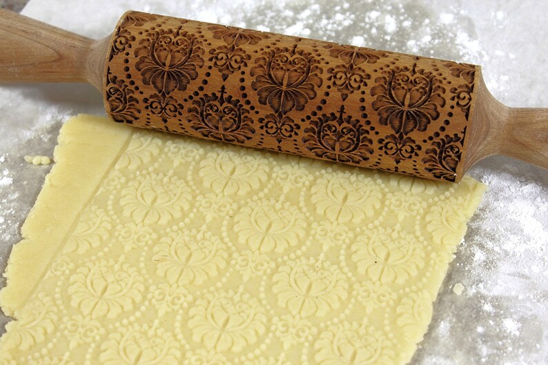 May include: A wooden rolling pin with an ornate floral pattern is pressed into a sheet of dough, creating an embossed design. The rolling pin is light brown, and the dough is a pale yellow color. Flour is sprinkled around the dough.
