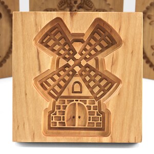 Puede incluir: Un cortador de galletas de madera con forma de molino de viento. El cortador est&aacute; hecho de madera marr&oacute;n claro y tiene un dise&ntilde;o detallado.