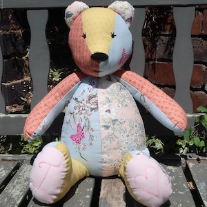 Pode incluir: Um urso de pelúcia patchwork feito à mão com um design colorido. O urso apresenta uma mistura de tecidos em tons de rosa, azul, amarelo e estampas florais. O urso está sentado em um banco de madeira desgastado.