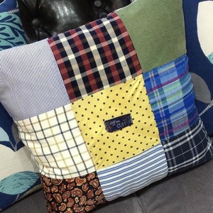 Może przedstawiać: Poduszka patchworkowa z różnymi tkaninami, w tym w kratkę, w paski i w wzory paisley. Poduszka jest żółta, niebieska, zielona i brązowa. Poduszka ma etykietę z napisem "Off the Cuff".