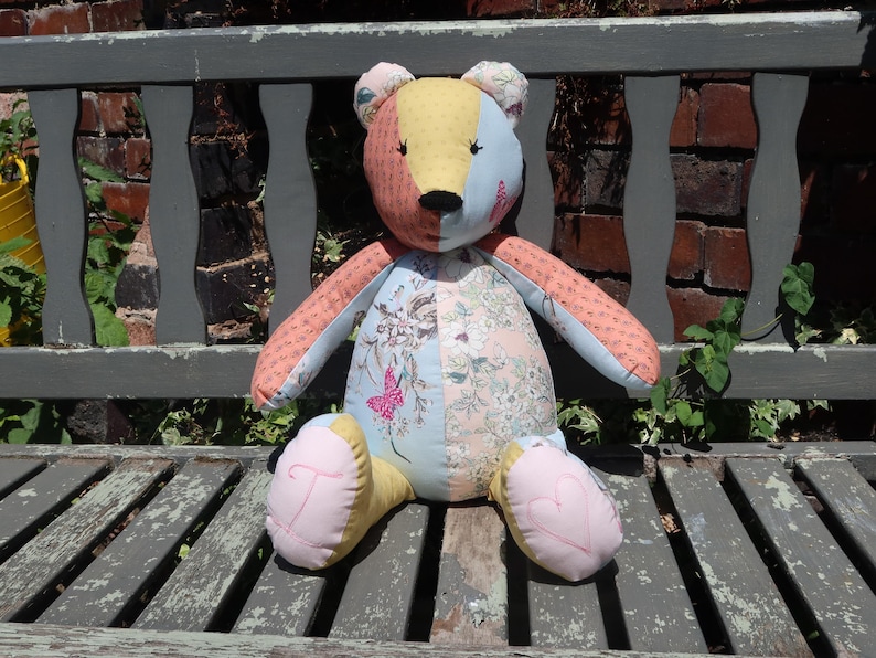May include: A handmade patchwork teddy bear with a colorful design. The bear features a mix of fabrics in shades of pink, blue, yellow, and floral patterns. The bear is sitting on a weathered wooden bench.