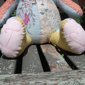 May include: A handmade stuffed animal with a patchwork design. The body features floral patterns and a butterfly. The feet are pink and yellow, with a stitched letter "T" and a heart. The toy is resting on a weathered wooden surface.