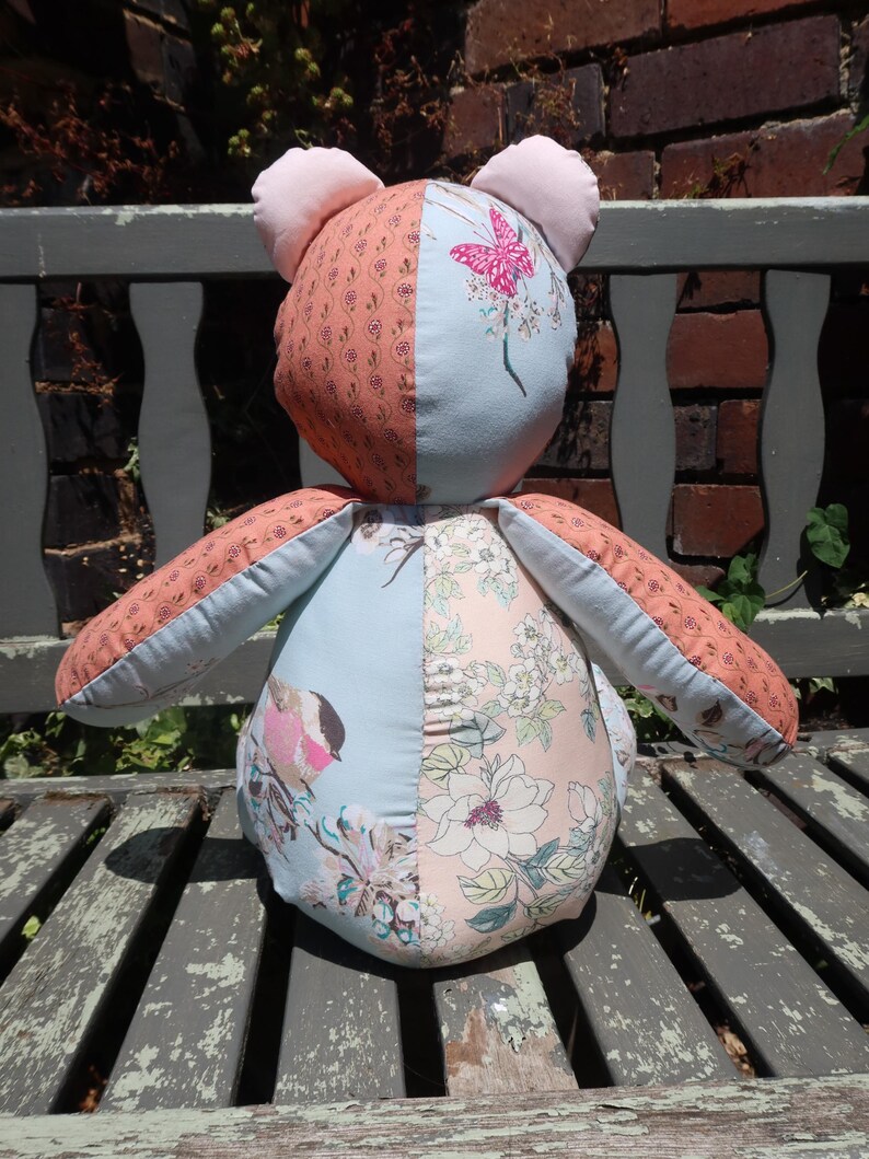 May include: A handmade stuffed bear with a patchwork design. The bear features a combination of floral and patterned fabrics in shades of pink, peach, and blue. The bear has pink ears and is sitting on a weathered wooden bench.