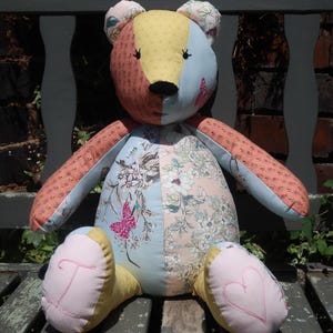 May include: A handmade patchwork teddy bear with a variety of floral and patterned fabrics in shades of pink, blue, yellow, and orange. The bear has embroidered details and is sitting on a wooden bench.