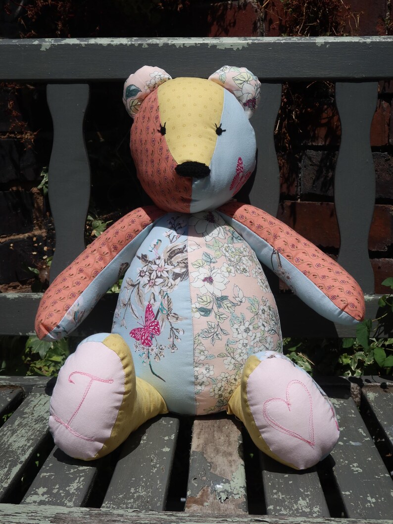 May include: A handmade patchwork teddy bear with a variety of floral and patterned fabrics in shades of pink, blue, yellow, and orange. The bear has embroidered details and is sitting on a wooden bench.