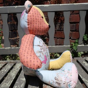 May include: A handmade patchwork teddy bear with a light blue body, peach arms, and yellow legs. The bear's head is a mix of peach, yellow, and light blue fabrics. Floral and butterfly patterns decorate the bear's body.