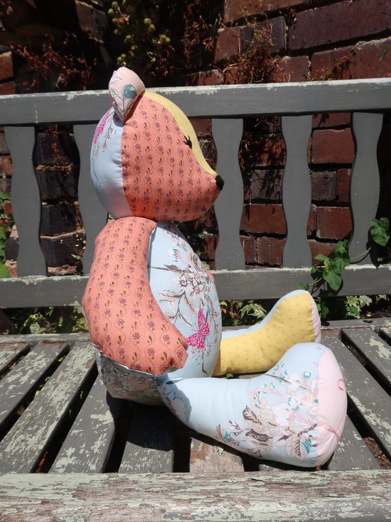 May include: A handmade patchwork teddy bear with a light blue body, peach arms, and yellow legs. The bear's head is a mix of peach, yellow, and light blue fabrics. Floral and butterfly patterns decorate the bear's body.