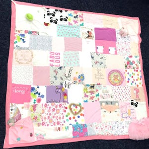 Op de afbeelding: Een patchwork quilt met een roze rand, bestaande uit vierkanten van verschillende stoffen. De vierkanten hebben prints van eenhoorns, panda's en bloemenpatronen. Sommige vierkanten hebben tekst zoals "FABULOUS" en "HAPPY!". De quilt is in roze en pastelkleuren.