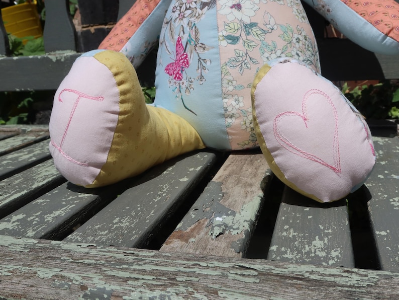May include: A handmade fabric doll with patchwork design. The doll's body is a mix of floral and solid fabrics in pastel colors. The doll's feet are pink and yellow, with a stitched letter "T" and a heart.