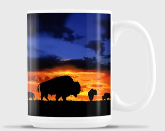 May include: White ceramic mug with a colorful sunset scene featuring a silhouette of a herd of bison. The mug has a large handle.