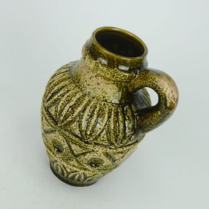 May include: A green ceramic vase with a textured, geometric design and a handle. The vase has a slightly distressed look, giving it a vintage feel.