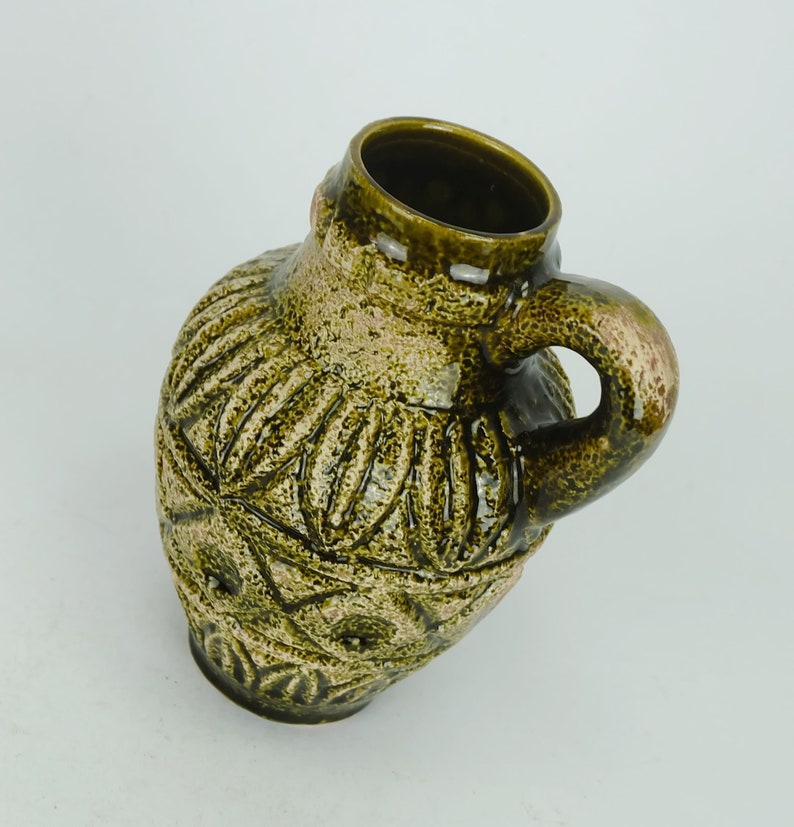 May include: A green ceramic vase with a textured, geometric design and a handle. The vase has a slightly distressed look, giving it a vintage feel.