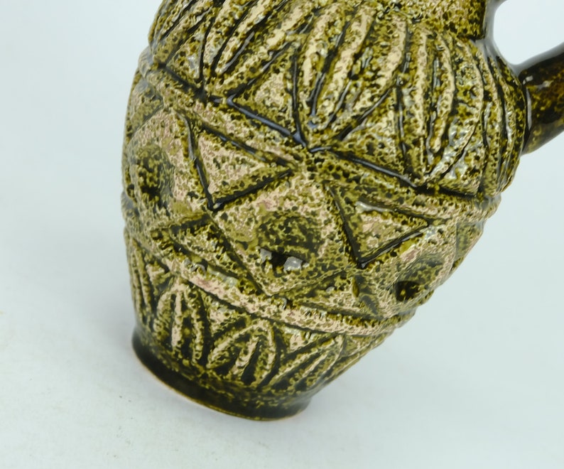 May include: A green ceramic pitcher with a geometric pattern. The pitcher has a textured surface and a wide mouth.