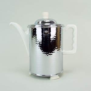 May include: A vintage silver-colored coffee pot with a white ceramic spout, handle, and lid knob. The pot has a hammered texture and a shiny, reflective surface. The lid is hinged and the pot sits on small white feet.