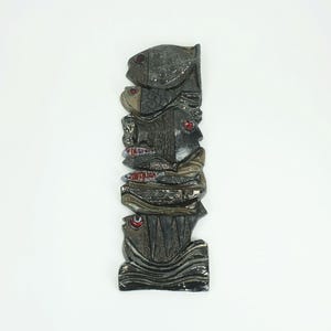 May include: A ceramic wall sculpture of a stack of fish with red eyes and a black, brown, and grey glaze. The fish are stacked on top of each other and appear to be swimming in a wave pattern.