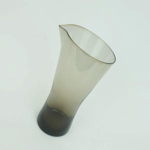 May include: A clear glass pitcher with a gray tint and a wide, flared opening. The pitcher has a smooth, rounded shape and a slender base.