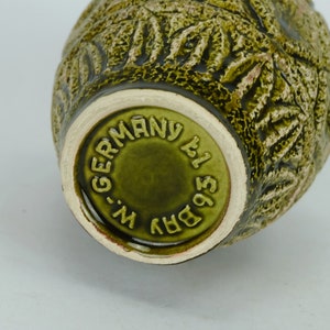 May include: Green ceramic vase with a textured surface and a raised design. The bottom of the vase has a circular stamp that reads "BRV 1951 W-GERMANY".