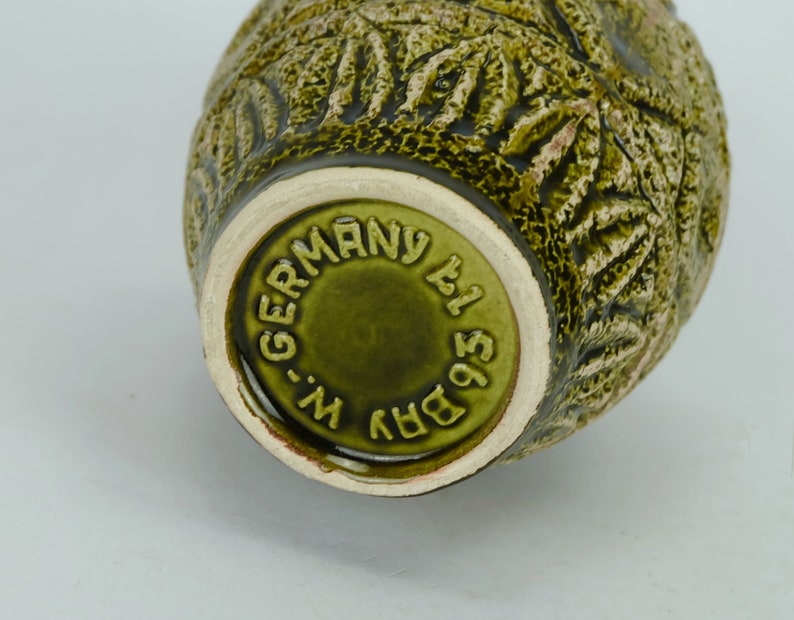 May include: Green ceramic vase with a textured surface and a raised design. The bottom of the vase has a circular stamp that reads "BRV 1951 W-GERMANY".