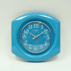 May include: A bright blue wall clock with a glossy finish and a rounded hexagonal frame. The clock face features white stylised numerals from 1 to 12, white hands, and the text Dubena Electric printed in white near the centre of the dial.