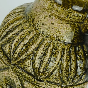 May include: A green ceramic pitcher with a textured surface and a handle. The pitcher has a unique design with a pattern of raised lines.