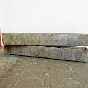 以下が含まれることがあります： 2つの風化した長方形のコンクリートプランター。プランターは積み重ねられており、垂直の溝があるテクスチャ加工された表面を示しています。色はくすんだグレーで、経年変化と屋外での使用を示唆しています。