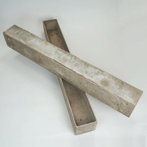 May include: Two rectangular, open-top concrete planters. The planters are a light gray color with a weathered appearance. They are long and narrow, suitable for planting flowers or herbs. The planters are placed on a white surface.