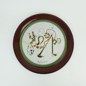 Peut inclure: Une assiette en céramique blanche avec un bord vert et marron. L'assiette présente un motif peint à la main de figures abstraites et de lignes dans des tons de vert, rouge, jaune et noir. La signature de l'artiste, "Porchier 80", est visible dans le coin inférieur droit.