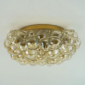 Pode incluir: Uma luminária de teto embutida com uma base circular de metal dourado e um conjunto de esferas de vidro transparente com tons âmbar dispostas em padrão de bolhas. Os globos de vidro variam em tamanho, criando uma superfície arredondada e texturizada.