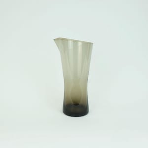 May include: A clear glass pitcher with a brown tint. The pitcher has a wide base and a narrow neck.