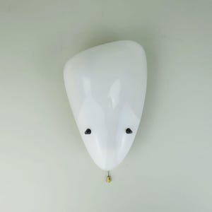 May include: White ceramic wall sconce with a rounded, organic shape. The sconce has two small black accents on the front and a gold-colored wire hanging from the bottom.