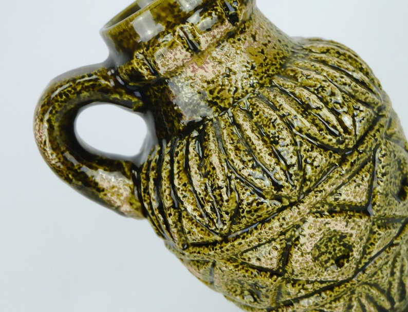 May include: A green ceramic vase with a textured surface and a geometric design. The vase has a wide mouth and a handle.