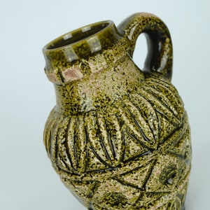 May include: A green ceramic pitcher with a textured surface and a handle. The pitcher has a unique design with a pattern of leaves and geometric shapes.