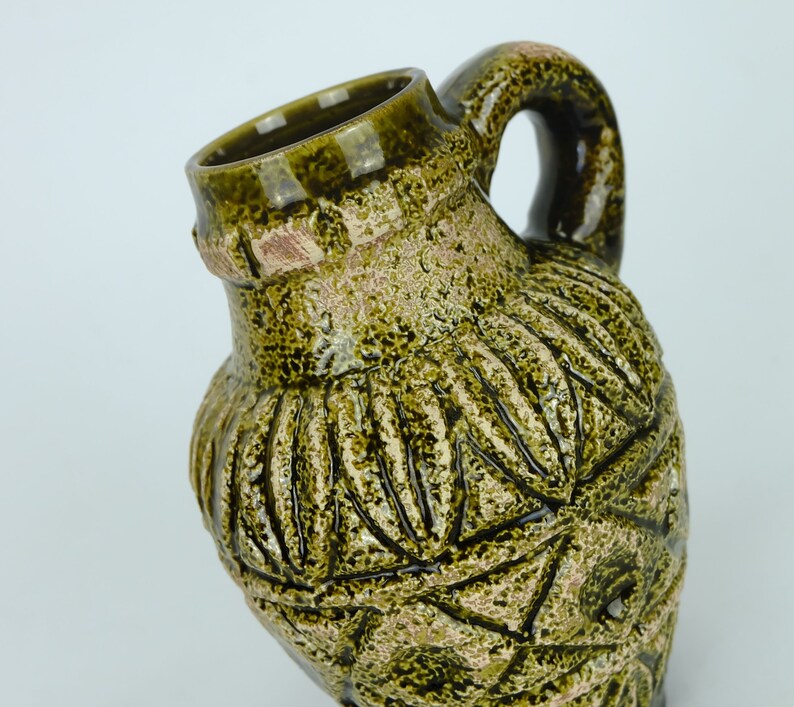 May include: A green ceramic pitcher with a textured surface and a handle. The pitcher has a unique design with a pattern of leaves and geometric shapes.
