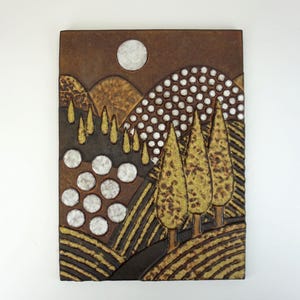 May include: A rectangular ceramic wall art piece depicting a landscape scene. The artwork features a brown background with a textured design, a full moon, rolling hills, and stylized trees in shades of gold and brown.