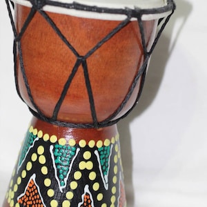 May include: A small brown wooden drum with a black rope net and a white top. The base of the drum is painted with a colorful geometric pattern of black, white, yellow, green, and orange.