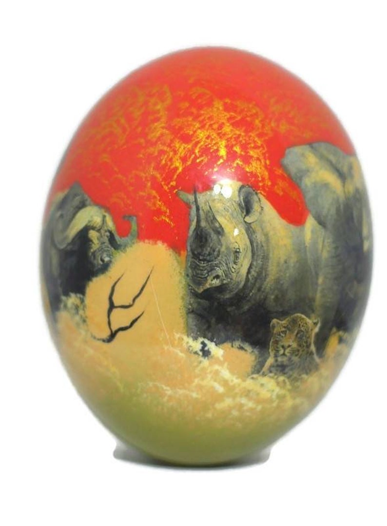 May include: A hand-painted ostrich egg with a red background and gold accents. The egg features a scene of a rhinoceros, a buffalo, and a leopard in a savanna setting.