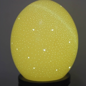 Puede incluir: Una lámpara de color amarillo en forma de huevo con una base blanca y un patrón de pequeños agujeros que emiten luz.