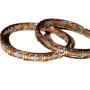 Könnte beinhalten: Zwei geflochtene Armbänder aus Silber, Kupfer und Gold. Die Armbänder sind ineinander verschlungen.