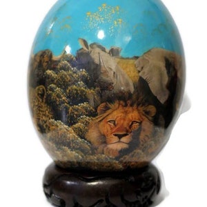 May include: Hand-painted ostrich egg with a scene of a lion, rhinoceros, and elephants in a savanna setting. The egg is displayed on a dark wood stand.