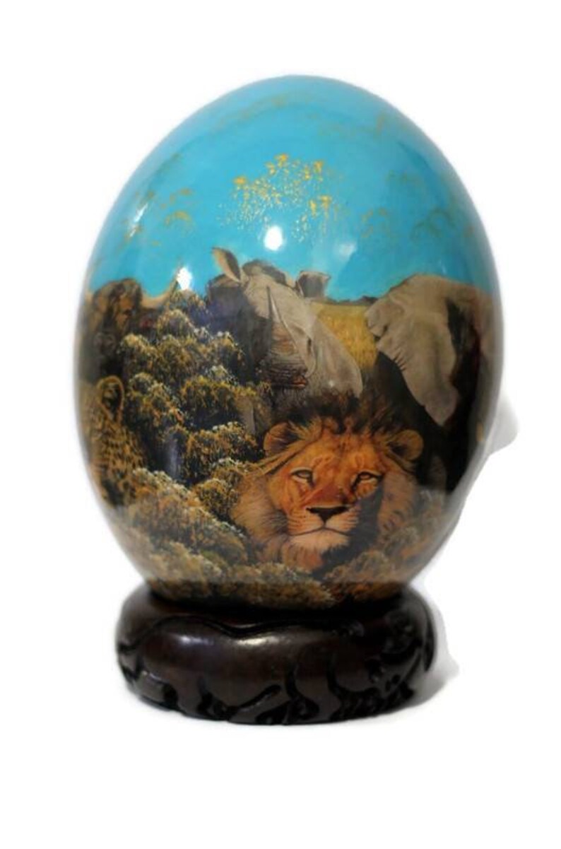 May include: Hand-painted ostrich egg with a scene of a lion, rhinoceros, and elephants in a savanna setting. The egg is displayed on a dark wood stand.