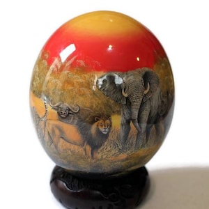 Può includere: Un uovo di struzzo dipinto a mano con una scena di fauna selvatica africana, tra cui un elefante, un leone e un bufalo, sullo sfondo di un tramonto. L'uovo è esposto su un supporto in legno.