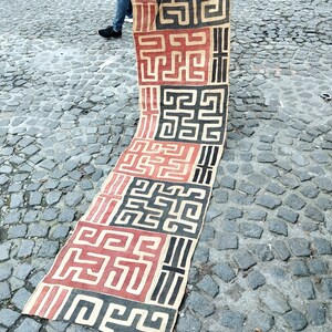 May include: A long, narrow, handwoven textile with a geometric pattern in black, red, and white. The textile is made of natural fibers and has a rough texture.