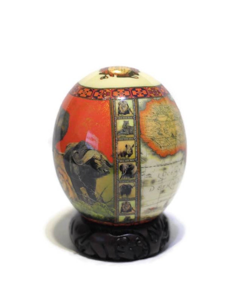 May include: A decorative ostrich egg with a map of Africa and images of African animals, including a lion, rhino, elephant, and buffalo. The egg is on a dark wood stand.