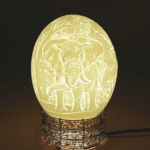 May include: A carved eggshell lamp featuring an elephant design. The eggshell is illuminated from within, showcasing the intricate details of the elephant and surrounding foliage. The lamp sits on a base of wrapped wire.